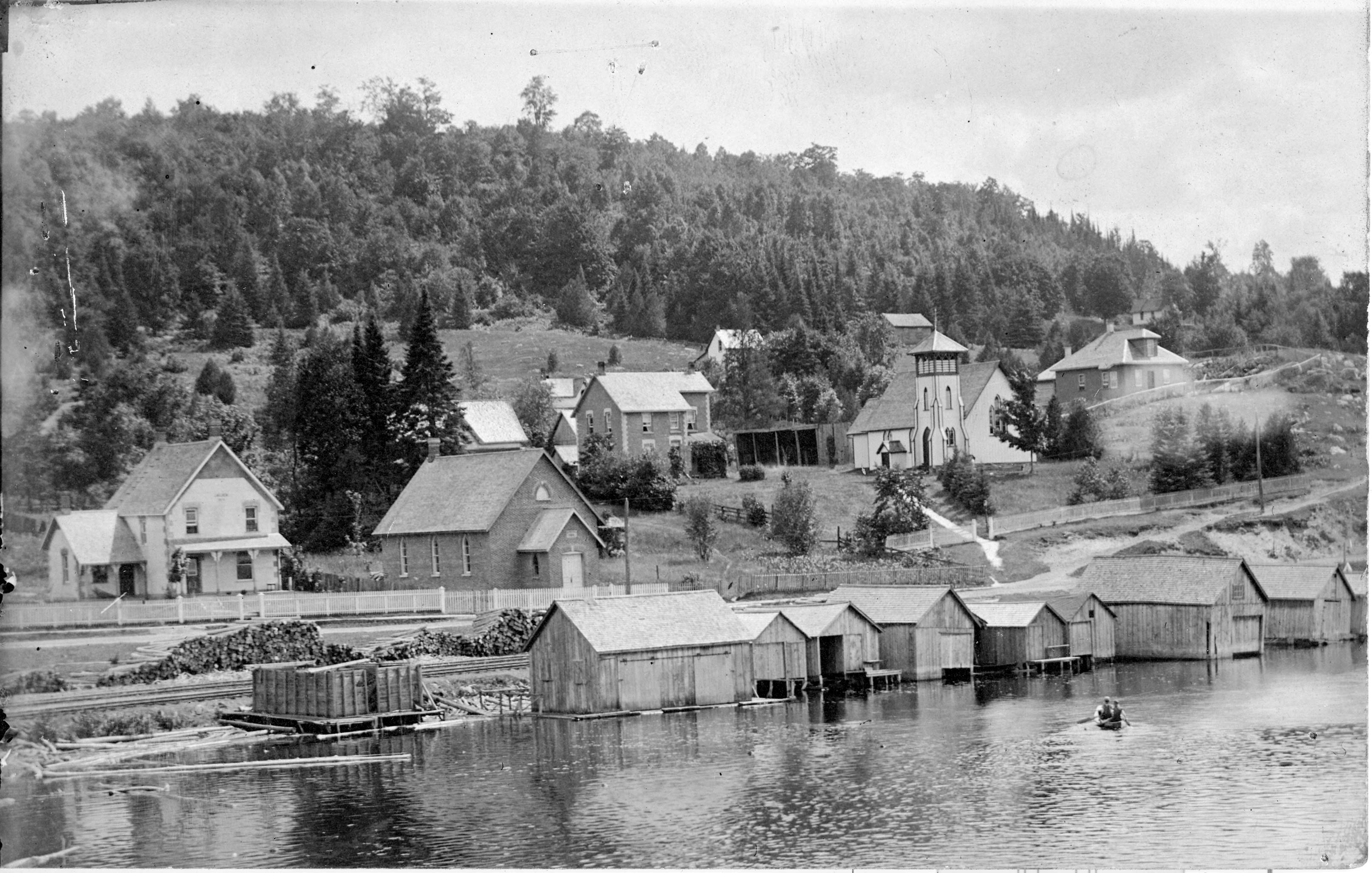 Picture looking at the shore of Head Lake. Along the shore there are boat houses, houses and towards the right back of the photo is a picture of the Anglican Church.